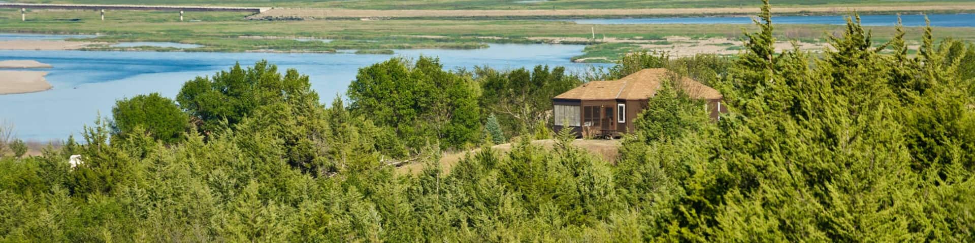 A house surrounded by dense forest, a river, and rolling hills in the background.
