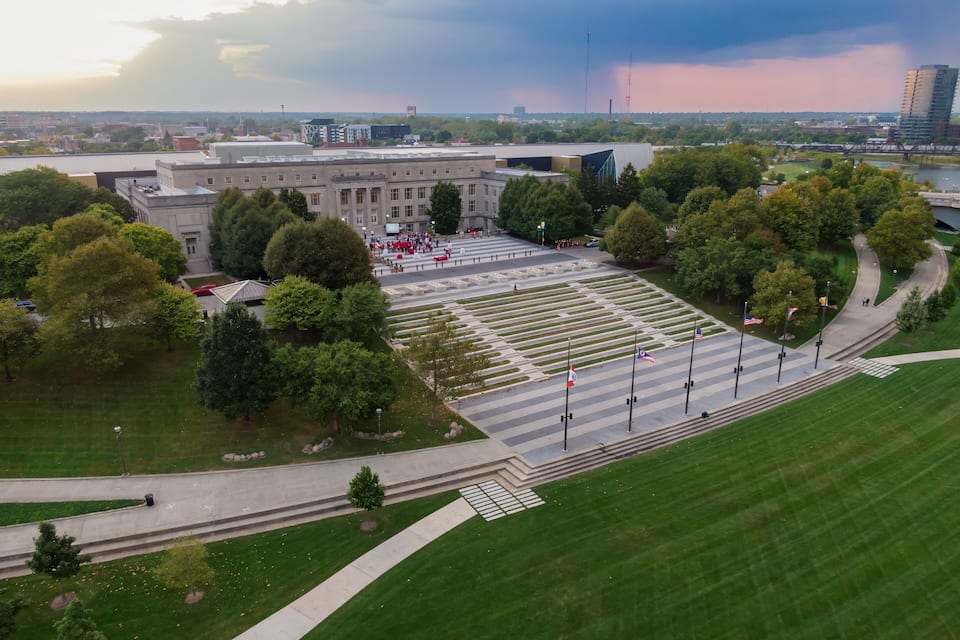 An aerial view of a memorial park with a large open plaza, flagpoles, and surrounding greenery.
