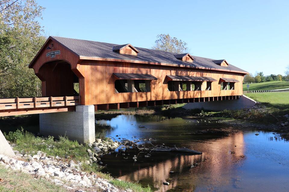 A wooden covered bridge over a calm river with a grassy bank and trees in the background.