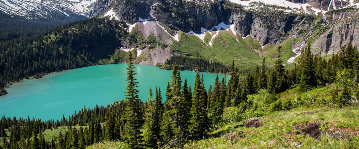 A turquoise lake surrounded by dense green forests and rugged mountains with snow-capped peaks.