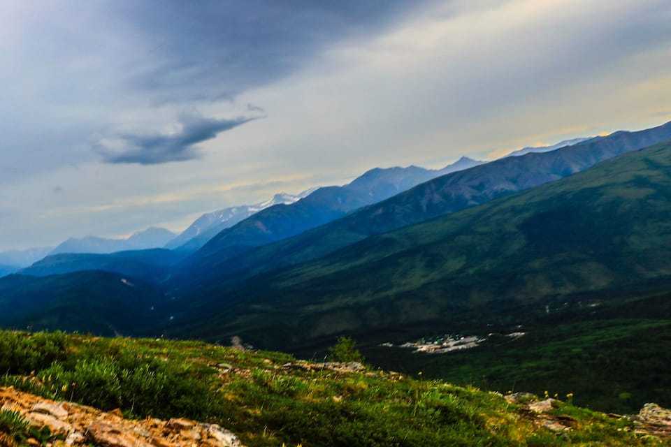 A mountainous landscape with a winding road, green hills, and distant snow-capped peaks under a cloudy sky.