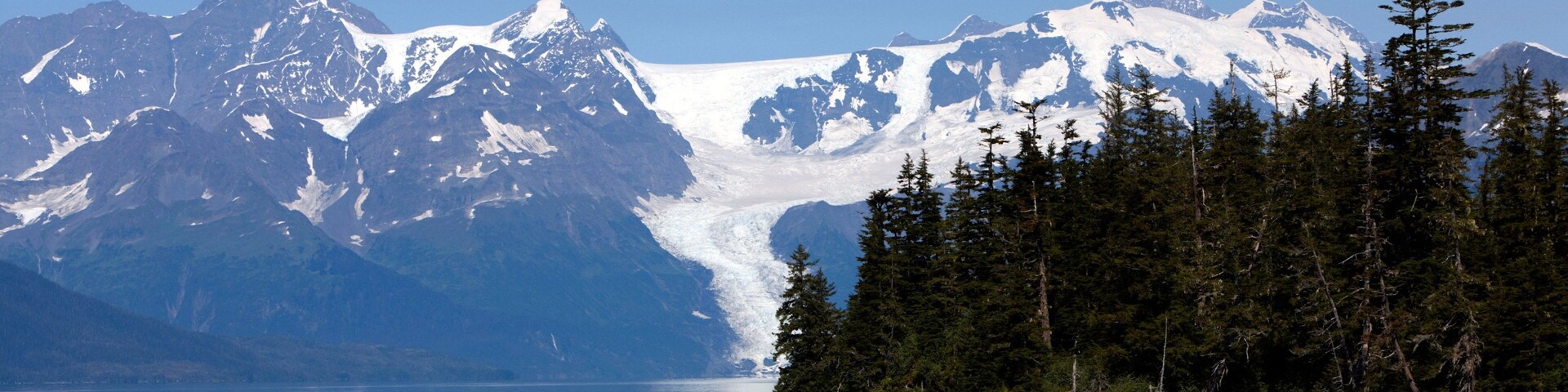 A glacier in front of snow-capped mountains, a calm lake, and a forested shoreline.
