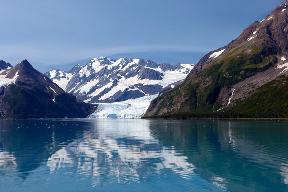 A glacier with a clear blue lake in the foreground and mountains in the background.