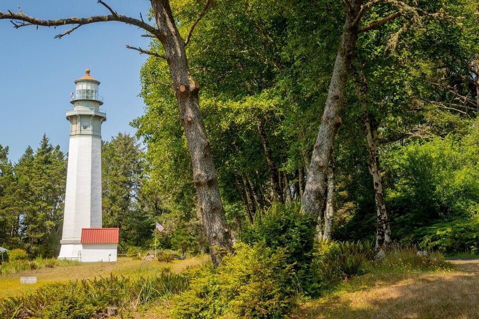 Valkoinen majakka, jossa on punainen katto, ja sen ympärillä on tiheää vihreää puustoa ja kirkas sininen taivas.