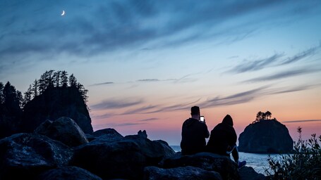 A rocky coastline at sunset with silhouettes of people and a crescent moon.