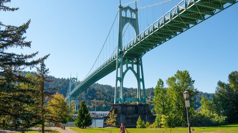 A large suspension bridge with a green structure, surrounded by a park with trees and a body of water in the background.