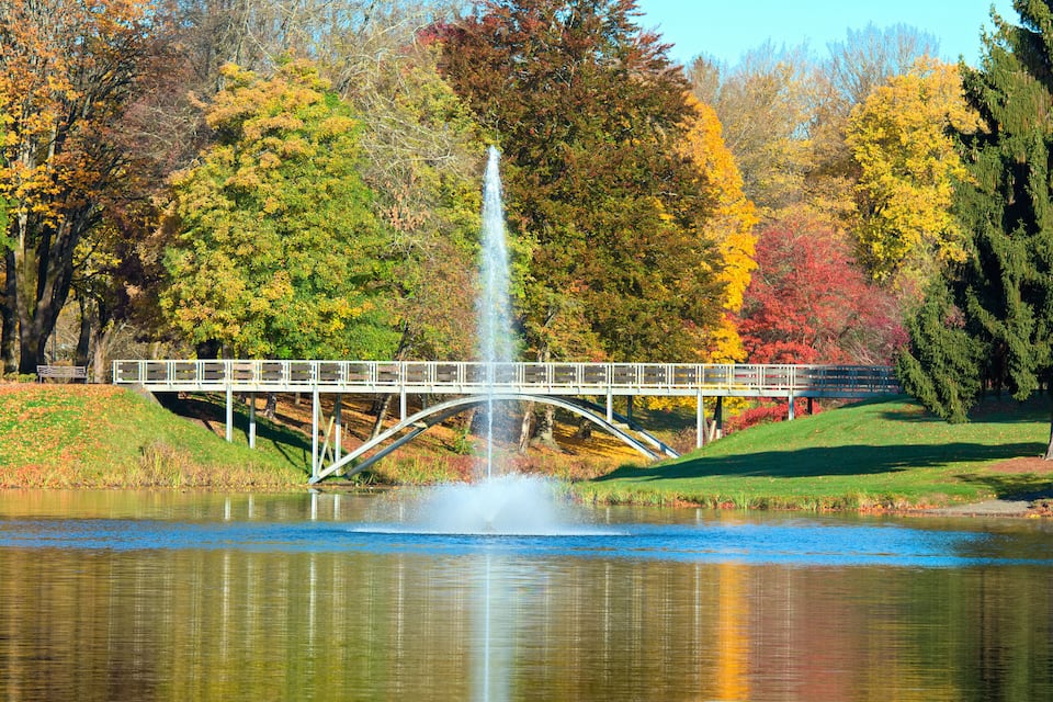 Un pont enjambant un étang orné d’une fontaine, le tout entouré d’arbres aux couleurs automnales.