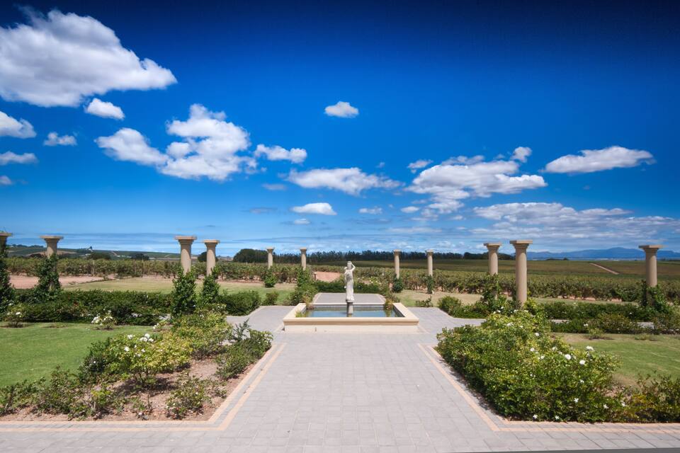 A landscaped garden with a central fountain, surrounded by columns and greenery under a clear blue sky.