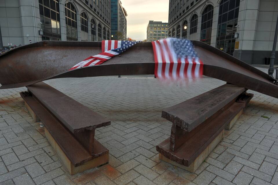 Una escultura de una estructura metálica curvada con banderas estadounidenses cubriéndola, ubicada en una plaza con bancos y edificios al fondo.