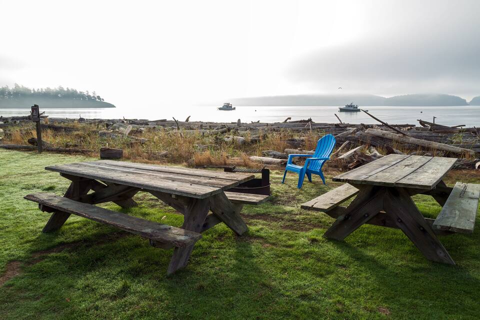 Un espace pique-nique avec des tables en bois et des chaises bleues, situé au bord d’un lac paisible, où l’on aperçoit des bateaux au loin.