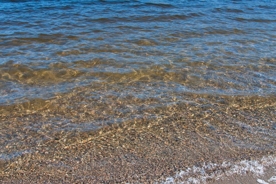 A lake with clear water, pebbly shore, and mountains in the background.