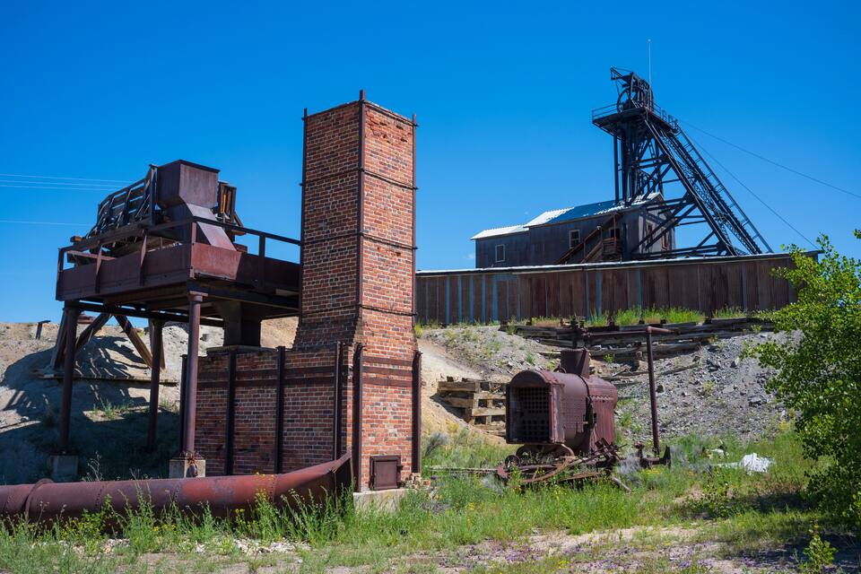 An abandoned industrial site with rusted machinery, a brick chimney, and a large metal structure.