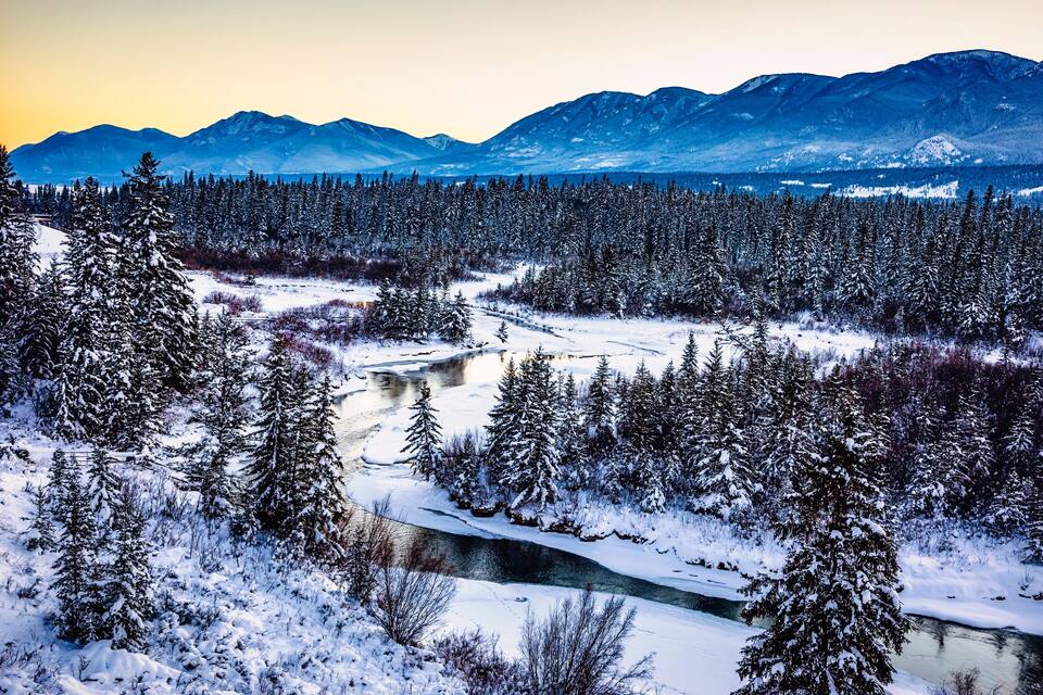 A snow-covered landscape with a river, dense coniferous forest, and mountains in the background.