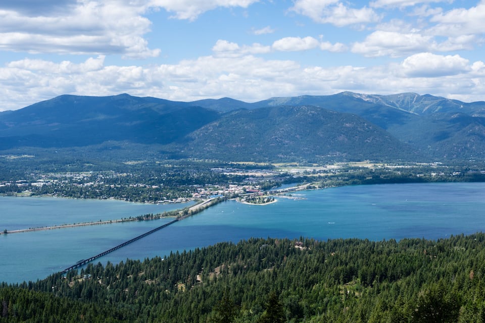 A coastal town with a pier extending into a large body of water, surrounded by mountains and dense forests.