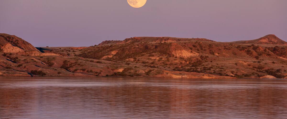 A moonlit landscape with a calm body of water reflecting the moon and surrounding hills.