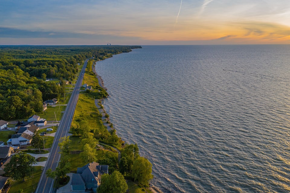 A coastal road with houses and greenery alongside a calm sea.