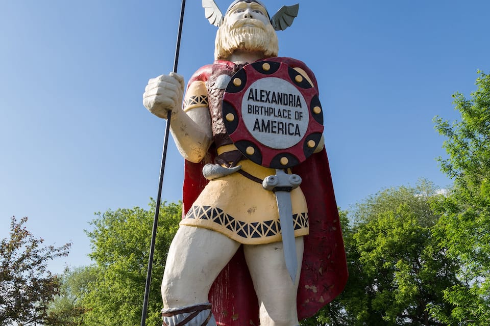 A statue of a warrior holding a spear, labeled "Alexandria Birthplace of America," set against a clear blue sky and green trees.