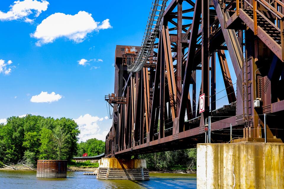 A large metal bridge with a chain and gears, supported by pillars in a river with trees and a clear sky.