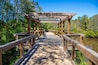 a long winding brown wooden bridge with a pergola over the Sandy Run Creek surrounded by lush green trees and plants with a gorgeous clear blue sky at The Walk at Sandy Run in Warner Robins Georgia