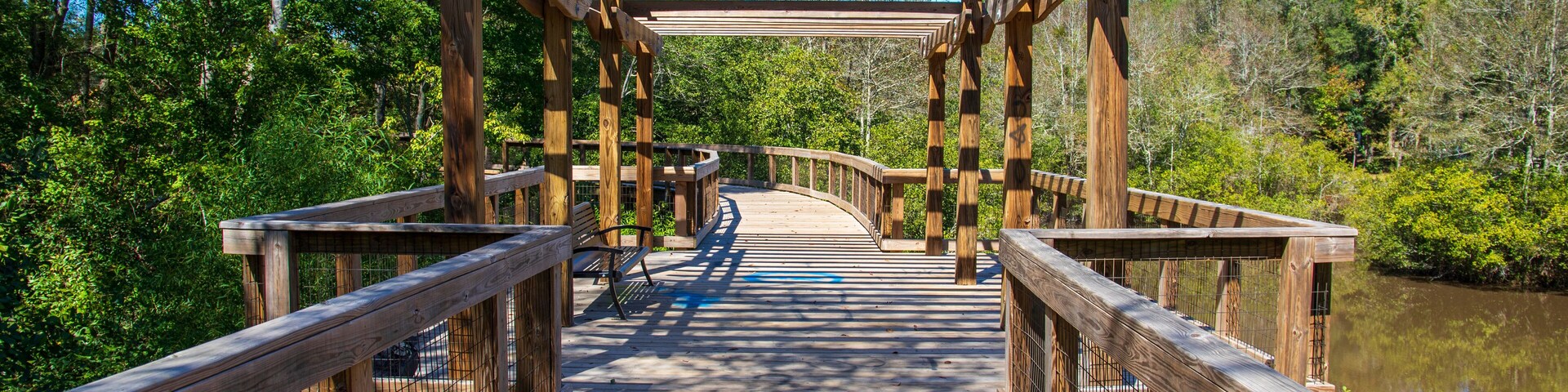 a long winding brown wooden bridge with a pergola over the Sandy Run Creek surrounded by lush green trees and plants with a gorgeous clear blue sky at The Walk at Sandy Run in Warner Robins Georgia