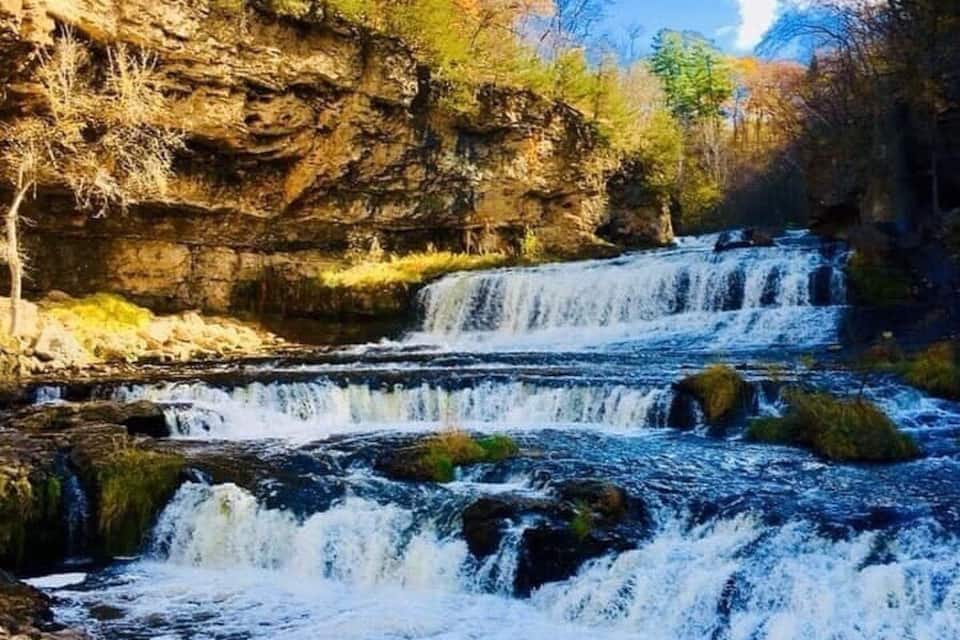 Une cascade à plusieurs niveaux, située dans une zone boisée, avec des falaises rocheuses et un feuillage automnal.