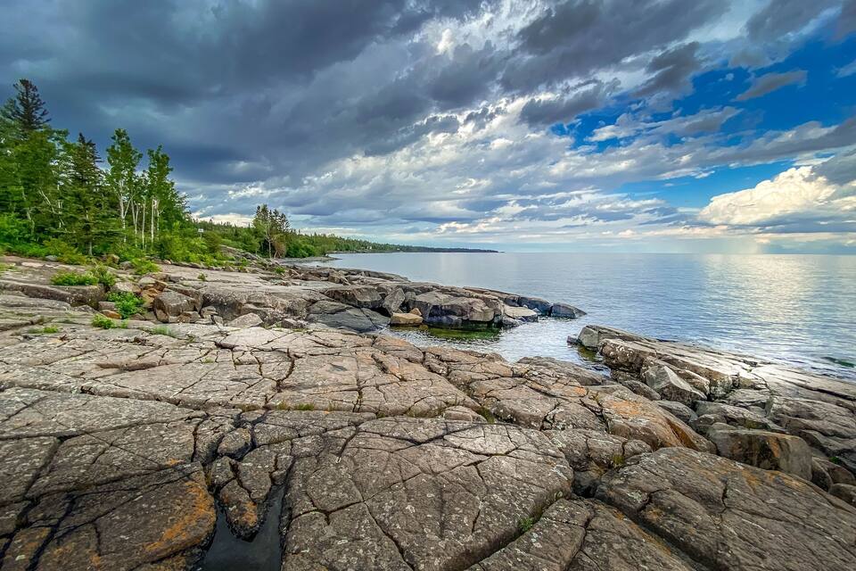 Une côte rocheuse bordant un lac paisible, entourée d’une forêt dense et verdoyante sous un ciel partiellement nuageux.
