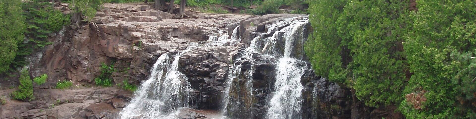 Una cascada de varios niveles desciende sobre terreno rocoso hacia un río tranquilo, rodeada de un denso bosque verde.