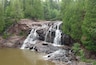 A multi-tiered waterfall cascading over rocky terrain into a calm river, surrounded by dense green forest.