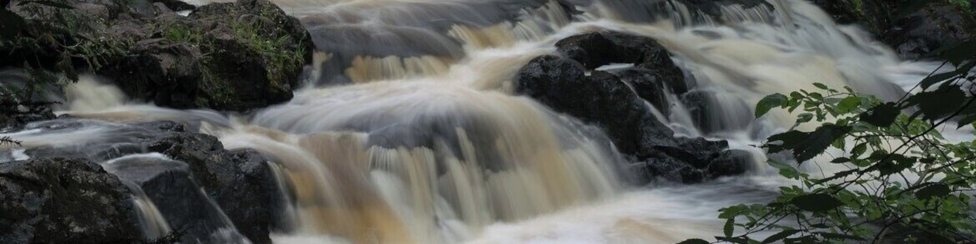 Ein schnell fließender Fluss mit mehreren Wasserfällen über Felsen, umgeben von dichtem Wald.