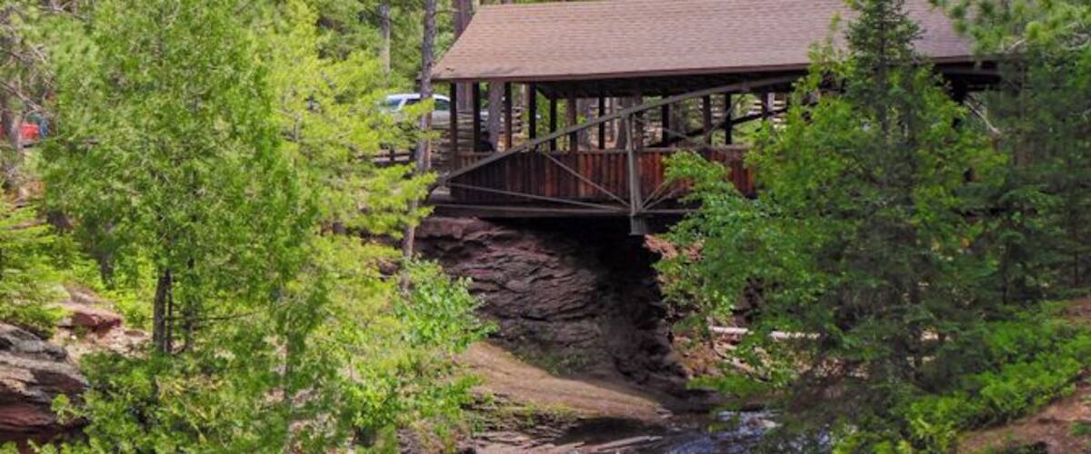 A wooden bridge over a river with a waterfall, surrounded by dense green trees.