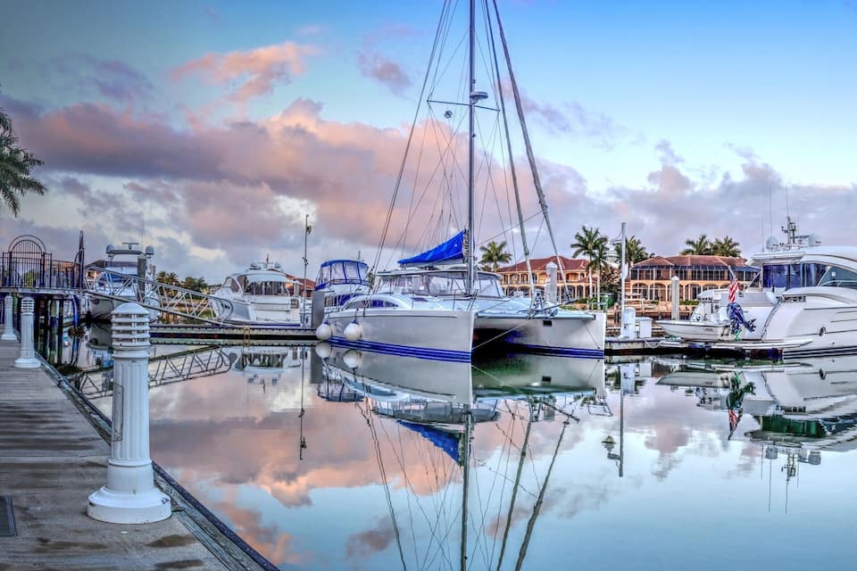 Un puerto deportivo con barcos amarrados, un paseo marítimo y edificios al fondo.