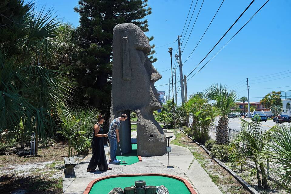 A large stone sculpture of a face in a park with palm trees and a green putting green.