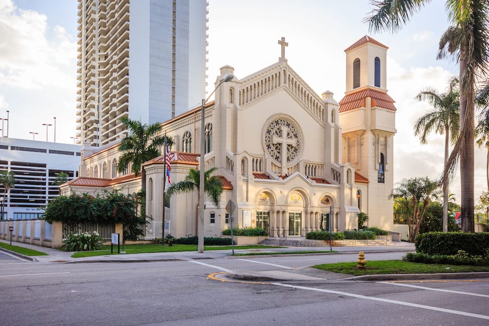 Une grande église dotée d’un clocher, entourée de palmiers et bordée d’un immeuble imposant.