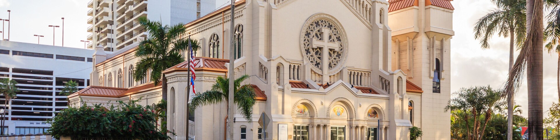 Una iglesia grande con un campanario, rodeada de palmeras y un edificio alto.