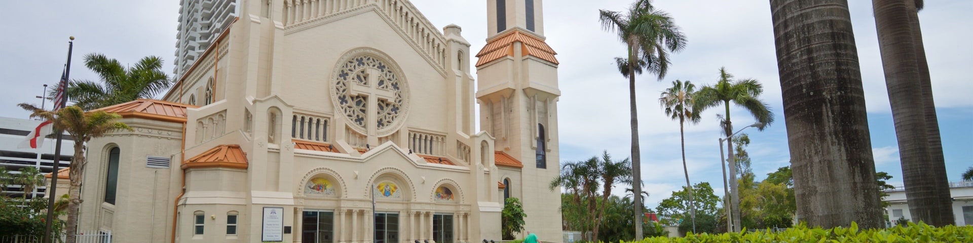 Una iglesia con una gran cruz, palmeras y un edificio alto al fondo.