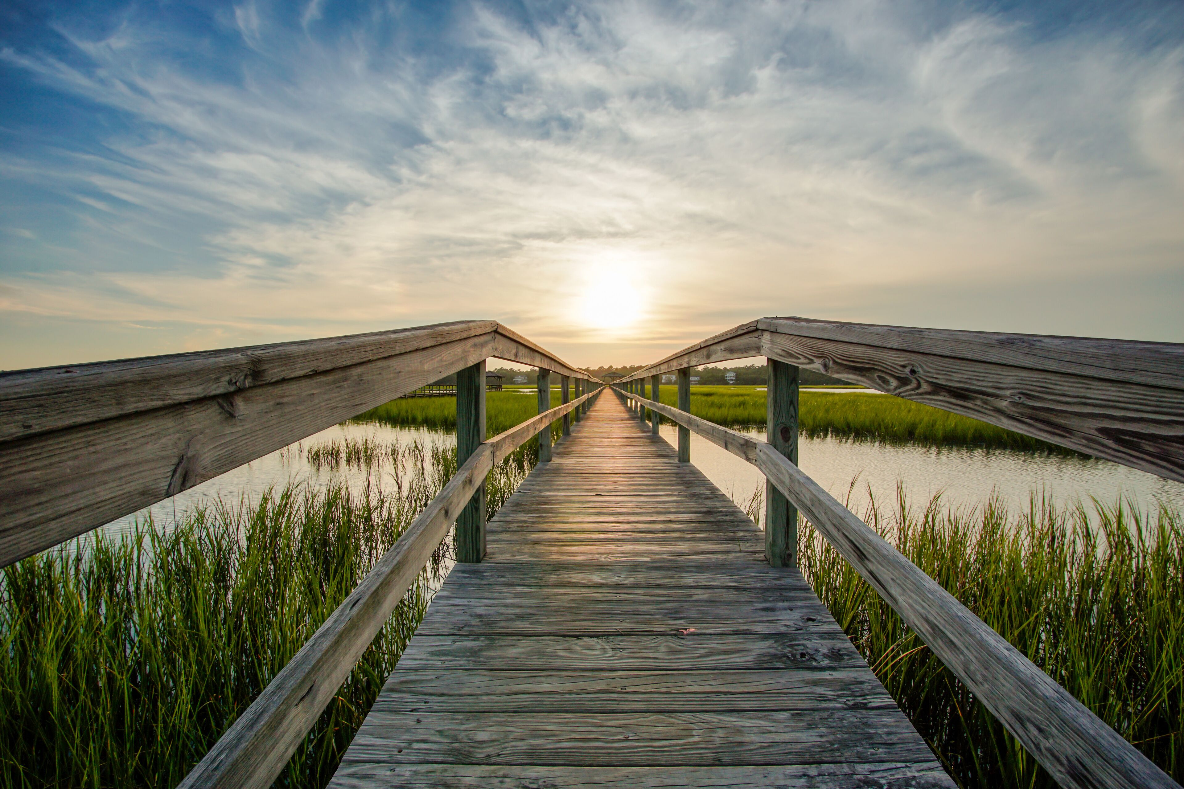 coastal waters with a very long wooden boardwalk pier in the center during a colorful summer sunset under an expressive sky with reflections in the water and marsh grass in the foreground