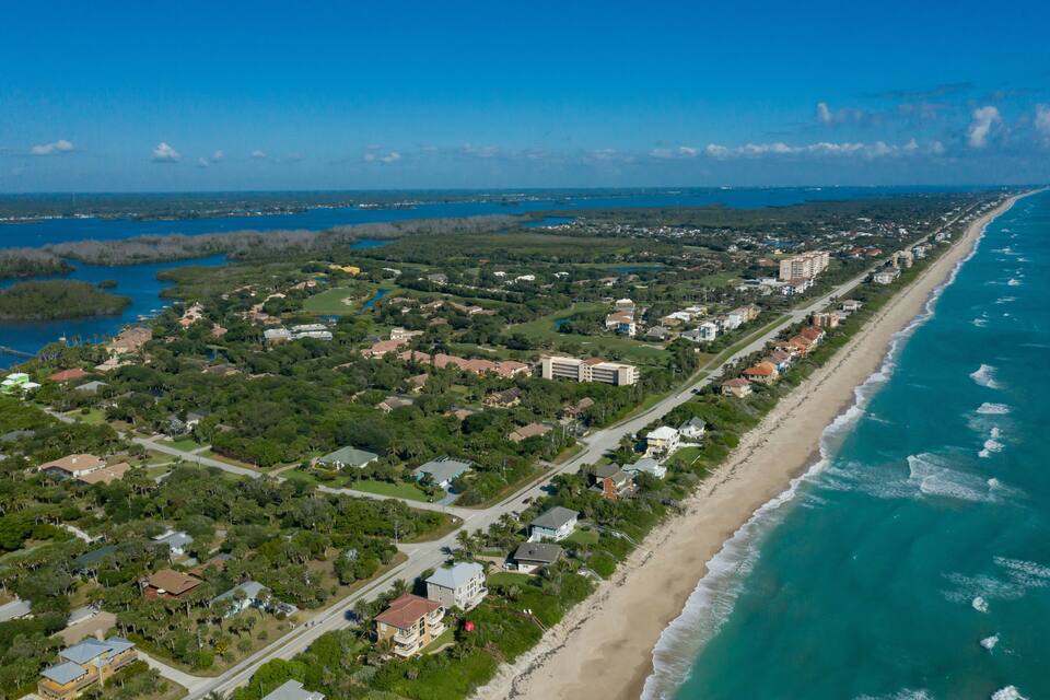 An aerial view of a coastal town with a long beach, dense greenery, and a clear blue sky.