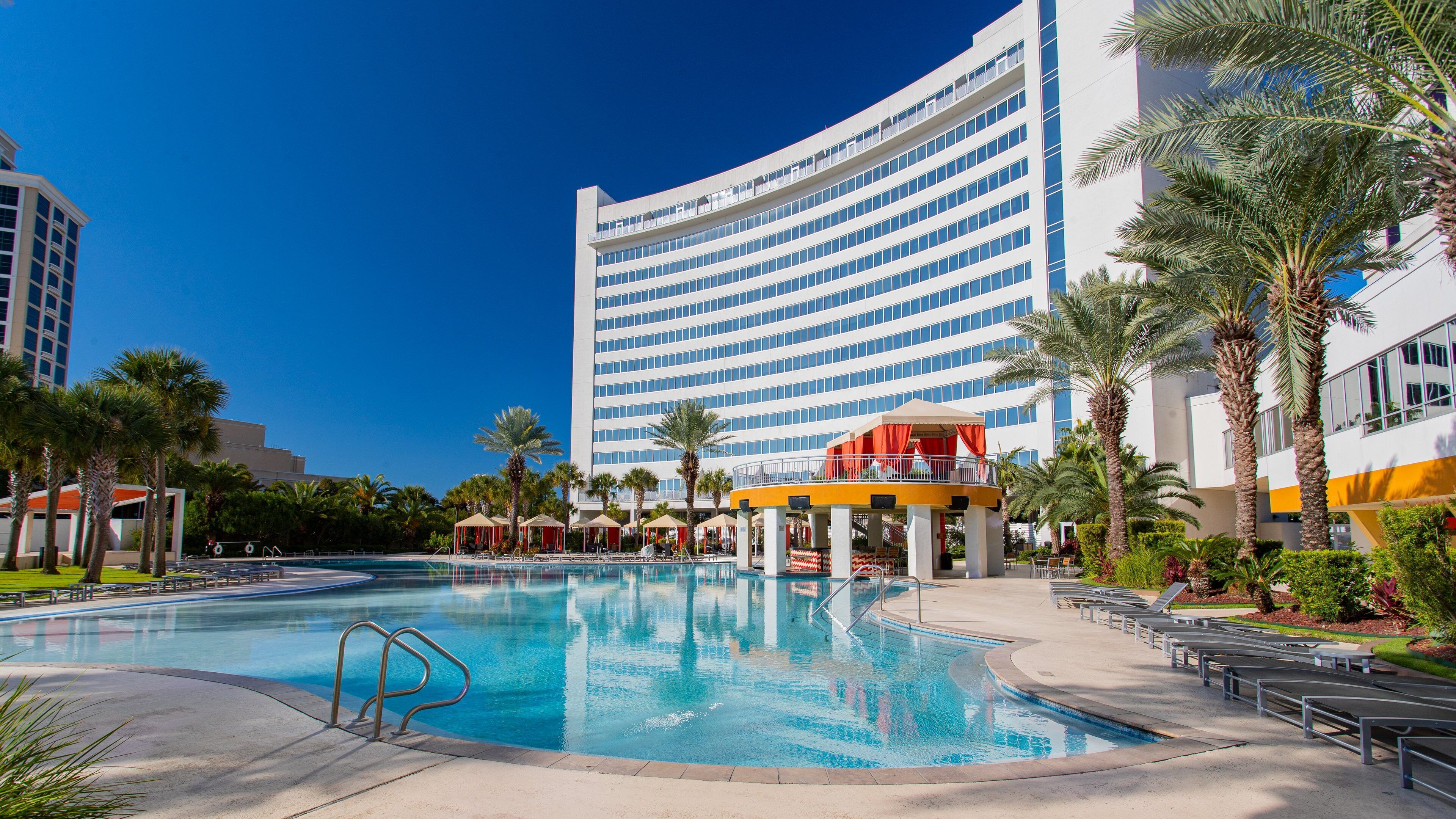 A swimming pool with lounge chairs and palm trees, surrounded by a modern building with a curved facade.