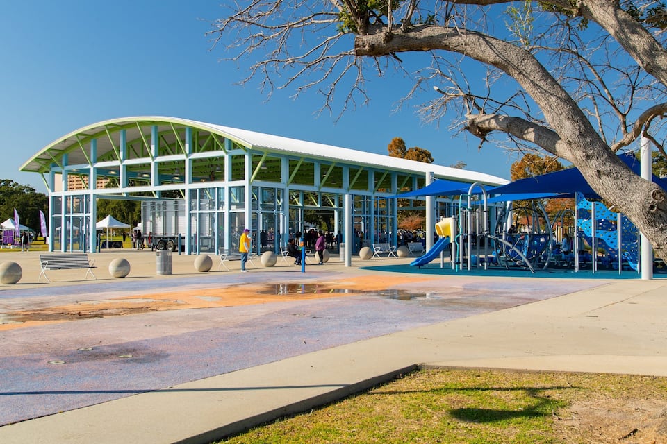A modern pavilion with a curved roof, surrounded by a playground with slides and climbing structures, and a tree in the foreground.