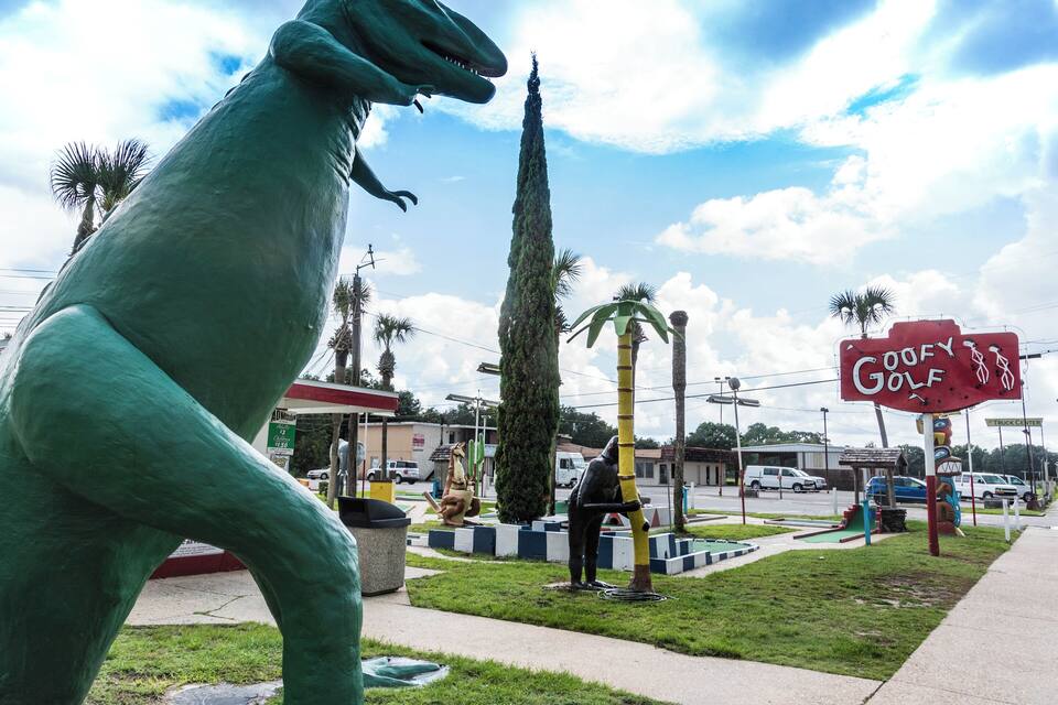 A dinosaur statue, a golf sign, and a tall tree in a park.