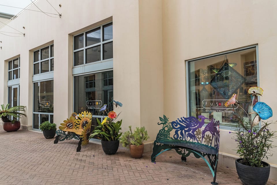 Un banco con diseños florales, plantas en macetas y una fachada con escaparates de vidrio.