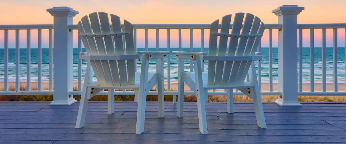 Zwei weiße Adirondack-Stühle auf einer Holzterrasse mit Blick auf den Ozean bei Sonnenuntergang.