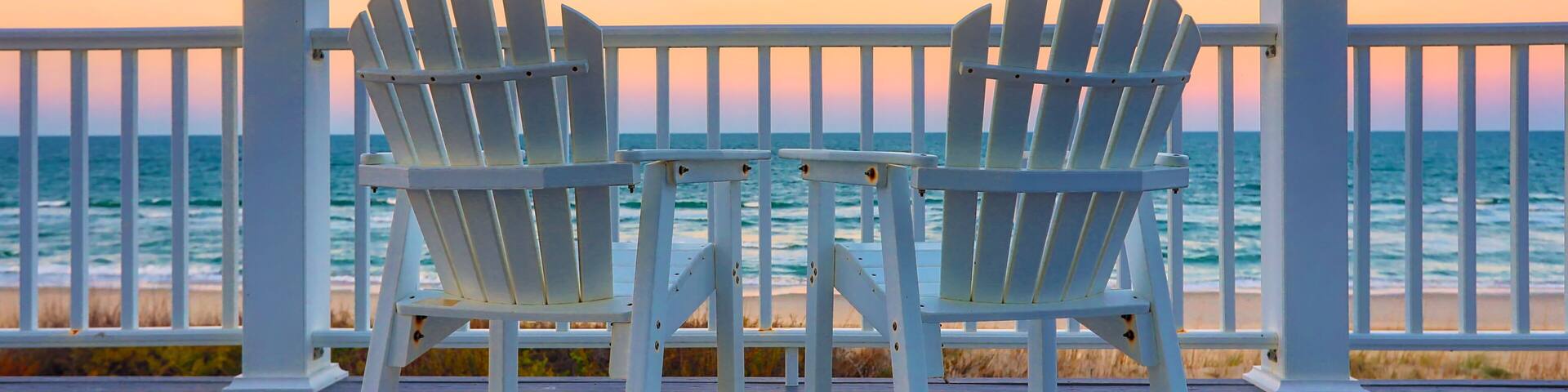 Two white Adirondack chairs on a wooden deck overlooking the ocean at sunset.