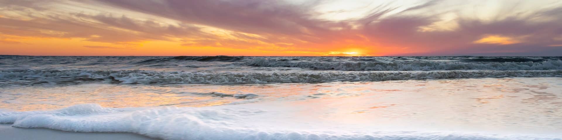Una playa con olas al atardecer, cielo despejado y el sol visible.