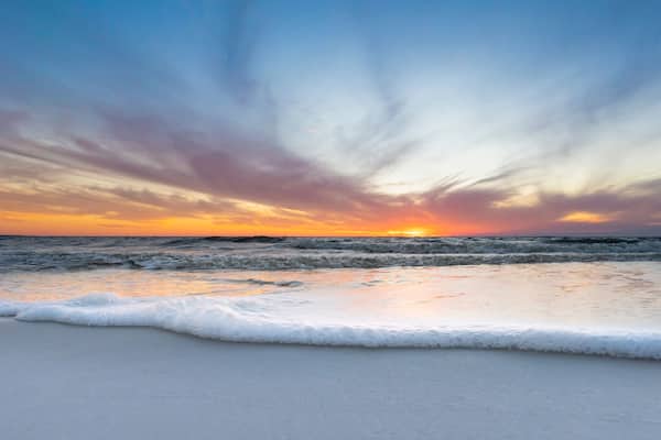 A beach with waves at sunset, a clear sky, and a visible sun.