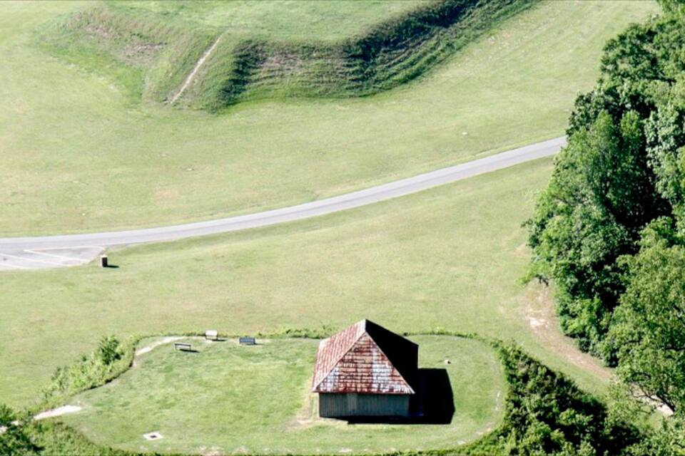 Ländliche Landschaft mit einem kleinen Haus, umzäunt, einer Straße und einem Hügel mit dichtem Baumbestand.