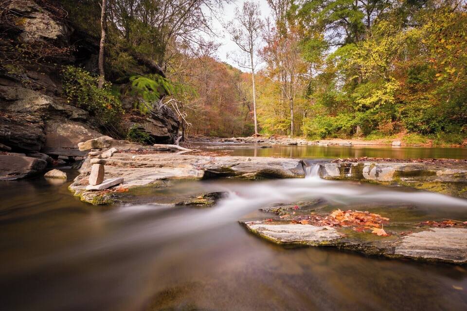Une rivière paisible, avec un lit rocheux, bordée d’un feuillage automnal et d’arbres.