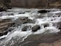 Une rivière aux eaux tumultueuses, où l’eau dévale en cascade sur les rochers, le tout étant entouré d’une zone boisée.
