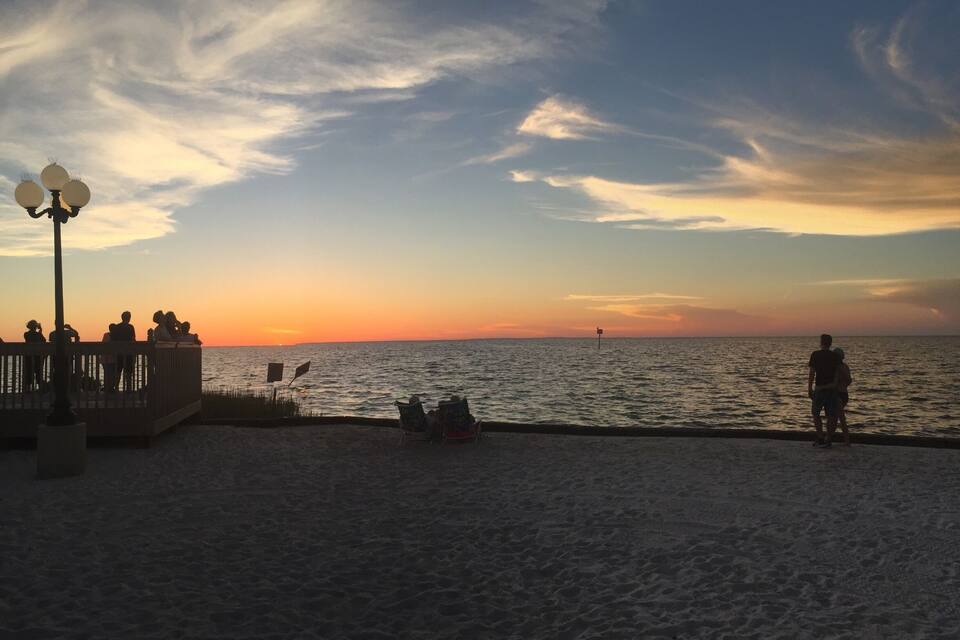 A beach at sunset with people sitting and standing on the sand, a lamp post, and a fence.