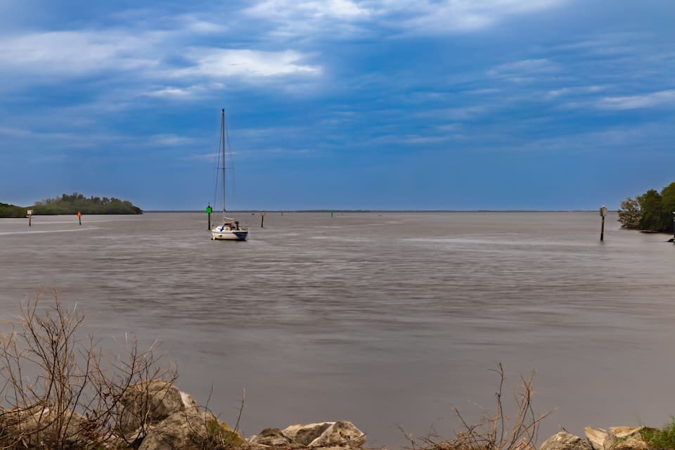 A calm body of water with a sailboat, a distant shoreline, and a clear sky.
